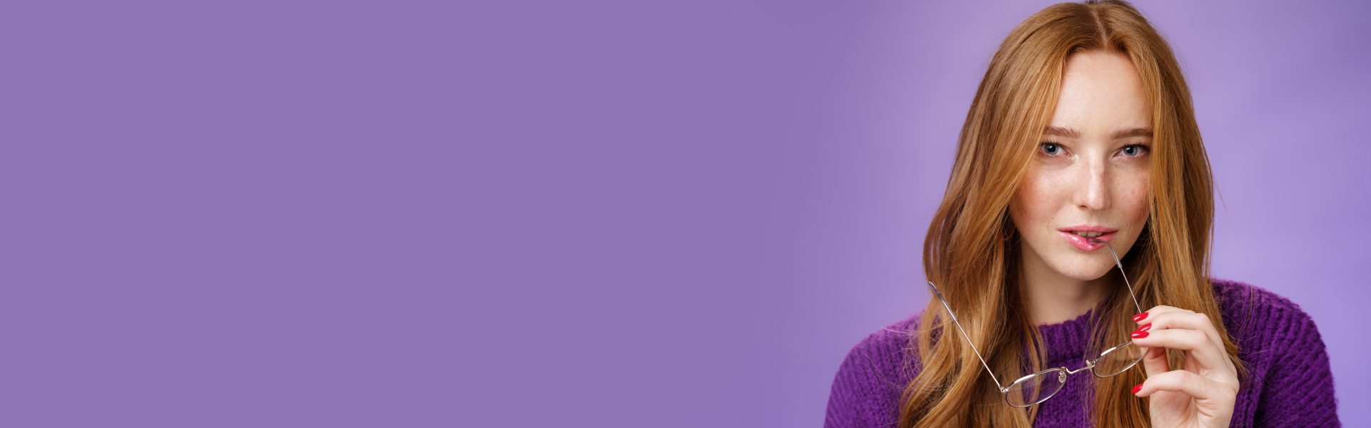 Close-up shot of smart and creative female redhead genius biting frame of glasses and squinting curious at camera as thinking having idea, making up plan against purple background.